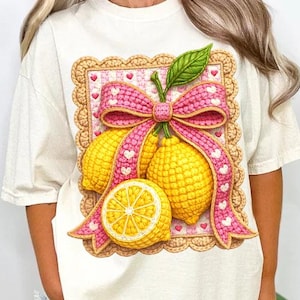 Puede incluir: Camiseta color crema con un gráfico de limones, un lazo rosa y un borde decorativo. Los limones son amarillos con una hoja verde, y el lazo tiene un patrón de corazones. El diseño está centrado en la parte delantera de la camiseta.