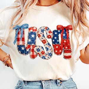 Puede incluir: Camiseta color crema con la palabra "USA" en un diseño de patchwork. Las letras están decoradas con patrones rojos, blancos y azules, incluyendo estrellas, flores y cuadros. Cada letra tiene un lazo decorativo.