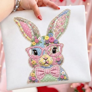 Puede incluir: Un cojín cuadrado blanco con un diseño de conejo patchwork. El conejo lleva gafas rosas y una pajarita, adornado con flores. El diseño incorpora patrones florales y colores pastel.