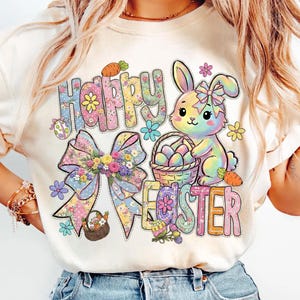 Puede incluir: Camiseta color crema con un gráfico de Pascua colorido. El diseño presenta las palabras "Happy Easter" en una fuente floral, un conejito arcoíris, una cesta de huevos, zanahorias y un gran lazo con flores.