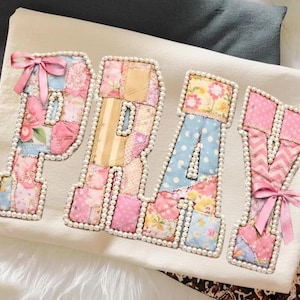 Puede incluir: Un artículo de tela blanca con la palabra "PRAY" aplicada en letras de patchwork. Cada letra está delineada con perlas de imitación y adornada con lazos rosas. El patchwork presenta patrones florales, a rayas y de lunares en colores pastel.