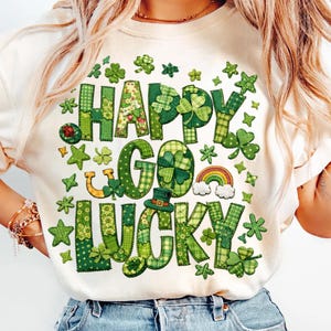 Puede incluir: Camiseta color crema con las palabras "HAPPY GO LUCKY" en letras verdes de patchwork. El diseño incluye tréboles, estrellas, un arcoíris y un sombrero de duende, todo en tonos verdes. Una camiseta festiva con temática del Día de San Patricio.