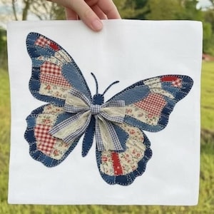Puede incluir: Un textil cuadrado blanco con una gran aplicación de mariposa. La mariposa tiene alas de patchwork en tonos azules, rojos y crema, con un lazo en el centro. Las alas están hechas de varias telas estampadas.