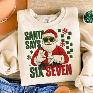 Puede incluir: Sudadera color crema con un gráfico de Papá Noel con gafas de sol y un traje rojo. El texto "SANTA SAYS SIX SEVEN" está impreso en verde y rojo. Copos de nieve y cuadrados verdes también aparecen en el diseño.