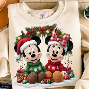 Puede incluir: Sudadera color crema con Mickey y Minnie Mouse con atuendo navideño. Mickey lleva un suéter verde y un gorro de Papá Noel, mientras que Minnie luce un suéter rojo y un lazo. Están rodeados de regalos, adornos y una corona festiva.
