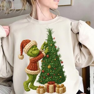 Puede incluir: Sudadera beige con un personaje de dibujos animados verde decorando un árbol de Navidad. El personaje lleva un atuendo y un gorro de punto rojo y blanco. El árbol está decorado con adornos y hay regalos en la base.