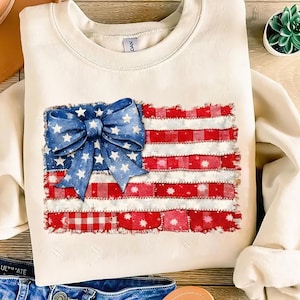 Puede incluir: Sudadera de color crema con un diseño patriótico. El diseño incluye un lazo azul con estrellas blancas y una bandera americana de patchwork en rojo y blanco. La sudadera tiene cuello redondo y mangas largas.