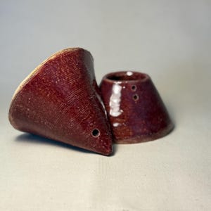 May include: Two handmade, maroon ceramic pieces. One is a cone-shaped pour-over coffee maker with a small hole near the tip. The other is a small, matching ceramic piece with two holes. Both have a textured surface.