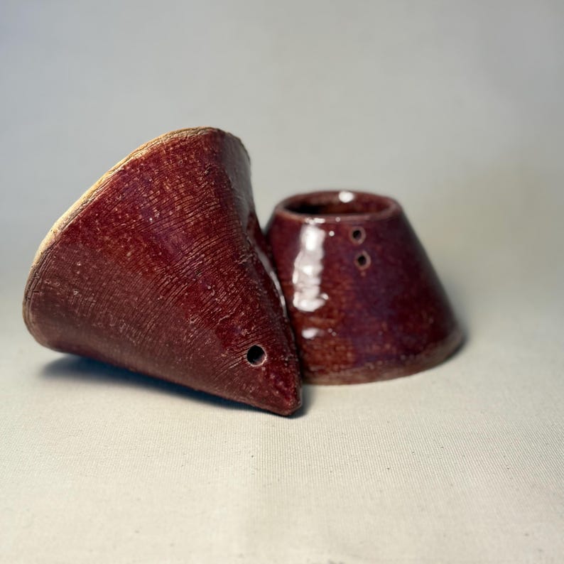 May include: Two handmade, maroon ceramic pieces. One is a cone-shaped pour-over coffee maker with a small hole near the tip. The other is a small, matching ceramic piece with two holes. Both have a textured surface.