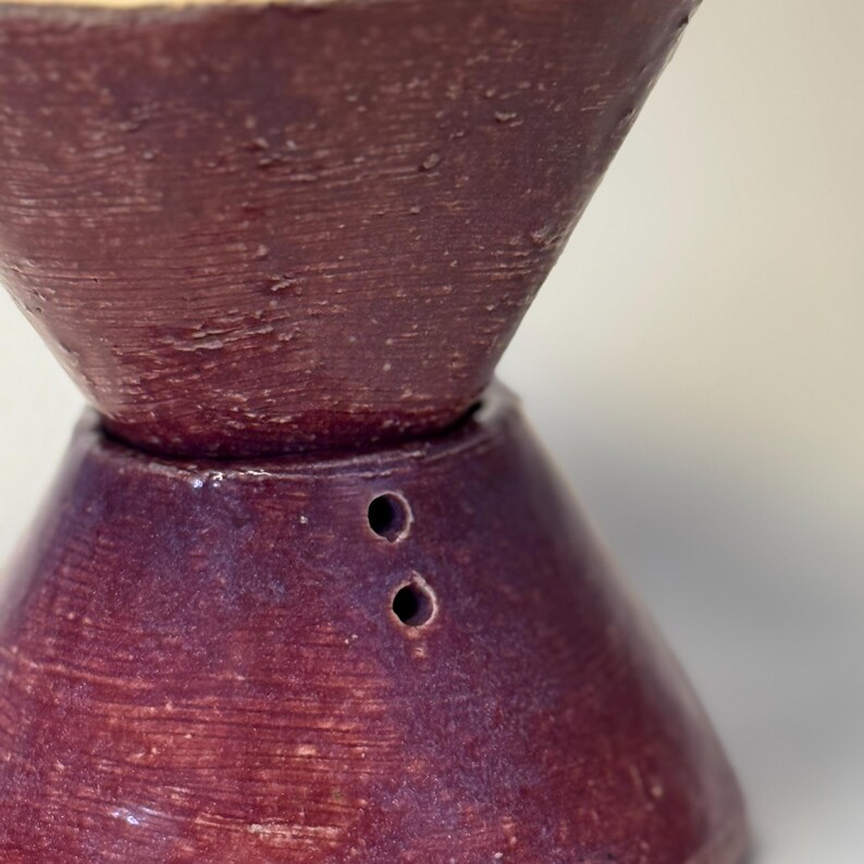May include: A close-up of a two-part, handmade ceramic pour-over coffee maker. The top section is a dark burgundy color, while the bottom section is a deep purple. Two small holes are visible on the bottom section. The surface has a textured, handmade appearance.