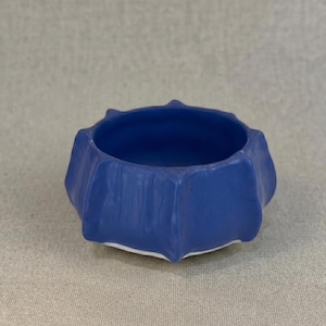 May include: A small, blue ceramic bowl with a distinctive, textured design. The bowl has a geometric shape with a white rim. The exterior is a matte blue, and the interior is a darker shade of blue.