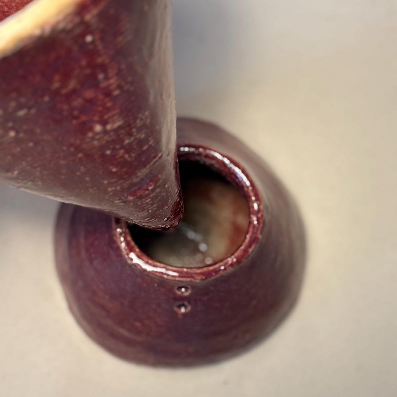 May include: A close-up of two handmade ceramic vases in a deep burgundy color. One vase is tilted, pouring into the other. The vases have a smooth, glossy finish and a unique, organic shape. The rim of the vase has a lighter color.