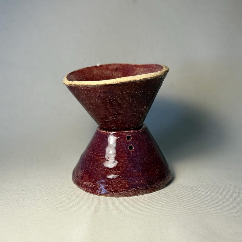 May include: A handmade, ceramic coffee pour-over with a deep burgundy glaze. The pour-over has a conical top and a flared base, connected by a short, cylindrical section with two small holes. The rim is a lighter, beige color.