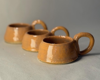 May include: Four handmade ceramic mugs in a warm, earthy orange-brown glaze. Each mug has a rounded body and a sturdy handle. The mugs are arranged in a row, with the closest one in sharp focus.