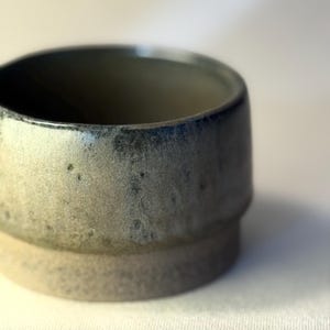 Op de afbeelding: Een handgemaakte keramische beker met een getextureerd, gespikkeld oppervlak. De beker heeft een tweekleurig kleurenschema, met een lichtbeige buitenkant en een donkerder, blauwgrijs interieur. De rand heeft een donkerblauwe glazuur. De beker heeft een getrapte basis.