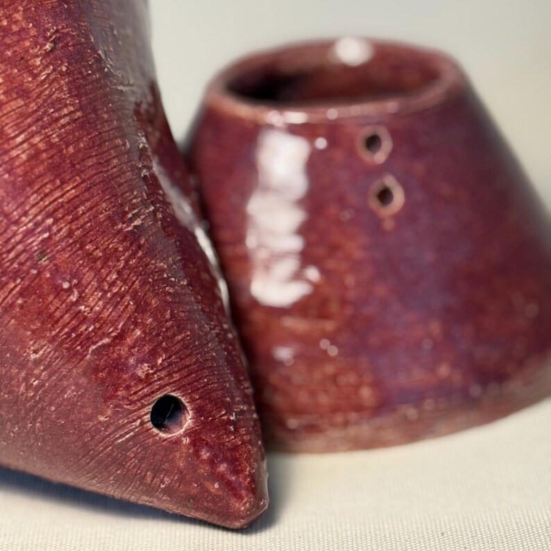 May include: Close-up of two maroon ceramic objects. One is a cone-shaped piece with a hole, and the other is a small, rounded cup with two holes. Both have a textured surface and a glossy finish.