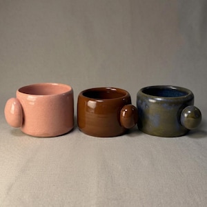 Pode incluir: Três canecas de cerâmica em fila, cada uma com uma cor única e uma alça arredondada. As canecas são rosa claro, castanho rico e azul-esverdeado com um acabamento salpicado. As alças têm uma cor semelhante ao corpo da caneca.