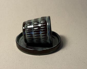 May include: A small, handmade ceramic cup and saucer set. The cup is dark brown with a textured, ribbed design and a blue glaze. The saucer is a matching dark brown with a simple, round shape. The set is ideal for serving espresso or small portions.