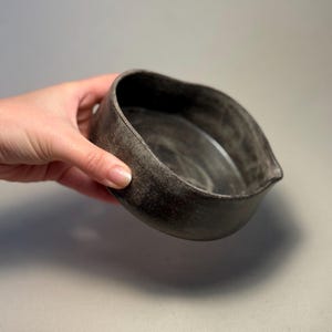 May include: A dark grey ceramic bowl with an unusual, asymmetrical form. The bowl has a textured surface and a small spout on one side. The bowl is held in a hand, showing its size and shape. The bowl's interior appears smooth, contrasting with the exterior.