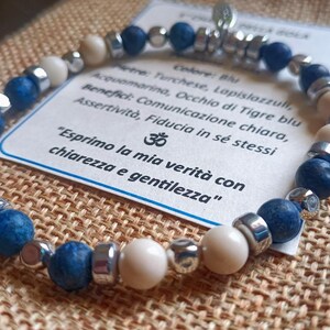 Può includere: Bracciale con perline blu, crema e argento. Il bracciale è esposto su una superficie beige strutturata. È visibile una piccola carta con testo in italiano. Le perline hanno un diametro di circa 0,8 cm.