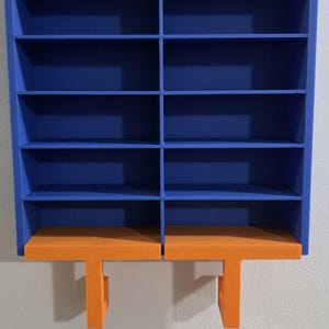May include: A blue wall-mounted storage organiser featuring ten rectangular compartments arranged in two vertical columns. The base of the unit includes two orange horizontal shelves, each supported by a vertical orange bracket extending downward.