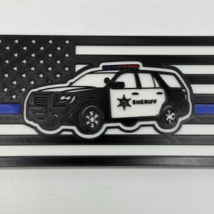 May include: A black and white 3D-printed American flag with a blue stripe and a sheriff's car silhouette. The car is black and white with the word "SHERIFF" and a star emblem on the side. The flag has a textured appearance.