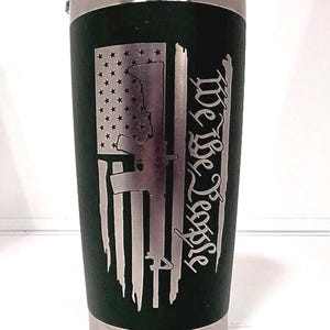 May include: A dark green insulated travel mug with a silver rim and lid. The mug features a distressed American flag design with a rifle silhouette and the words "We The People".