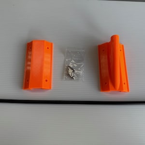 May include: An overhead shot shows components of a product kit. Two bright orange plastic pieces, a clear bag of hardware, and a long black rod are arranged on a white surface. The orange pieces have a rectangular shape with mounting holes.