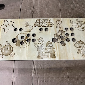Può includere: Pannello di gioco arcade in legno con personaggi di Super Mario incisi al laser e il logo del gioco. Il pannello ha fori preforati per pulsanti e joystick. Il design include Mario, Luigi, la Principessa Peach e altri personaggi iconici.