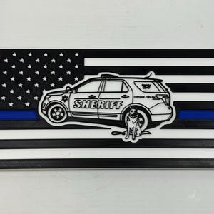 May include: A black and white American flag with a blue stripe and a white outline of a sheriff's vehicle and a dog. The word "SHERIFF" is printed on the side of the vehicle. The flag has a 3D printed appearance.