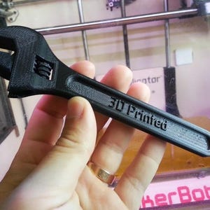 May include: Black adjustable wrench with the text "3D Printed" on the handle. The wrench is held in a hand and appears to be made of plastic. The wrench is a tool used for tightening or loosening nuts and bolts.