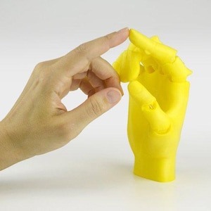 Może przedstawiać: Jasnożółta proteza dłoni jest pokazana z ludzką dłonią wchodzącą z nią w interakcję. Proteza dłoni jest wykonana z litego materiału i ma żywy żółty kolor. Ludzka dłoń wchodzi w interakcję z protezą dłoni.