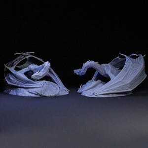 May include: Two light blue dragon figurines, each with detailed wings and a textured surface. One dragon is curled, while the other is posed with its wings spread. The background is a dark, solid color.