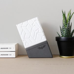 Puede incluir: Una caja bicolor con un patrón geométrico y un logotipo de Batman, junto a una planta suculenta en maceta y dos libros. La caja es blanca y gris, con un diseño texturizado en la mitad superior. Los libros son blancos con texto negro.