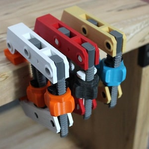 May include: Three colorful clamps, in white, red, and yellow, are attached to a wooden surface. Each clamp has a screw mechanism and is made of plastic. The clamps are used for woodworking or DIY projects.