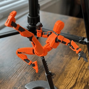 May include: An orange articulated action figure in a dynamic pose, with black accents on the joints. The figure is mounted on a black stand, set against a blurred background with a wooden surface.