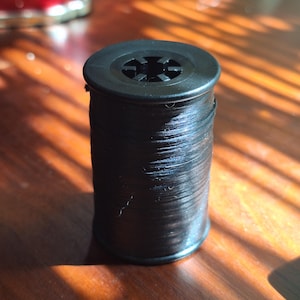 May include: A black spool of thread, tightly wound, with a plastic top featuring a star-shaped design. The thread is glossy and appears to be new. The spool is cylindrical and sits on a wooden surface.