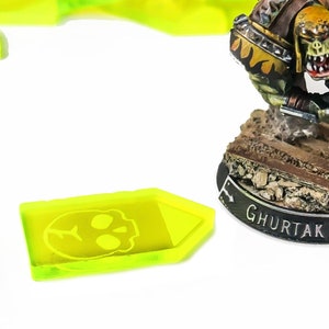 May include: A green and brown miniature figure of an Orc holding a gun, standing on a black base with the text "Ghurtar Gulggro". The figure is on a white background with a yellow plastic token.
