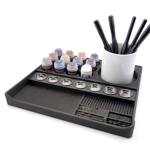May include: A black paint station with a variety of Citadel paint pots, small metal paint wells, and a white ceramic cup holding black paint brushes. The paint station has a textured surface and a small compartment for tools.
