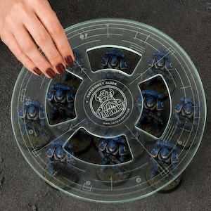 May include: A circular, transparent acrylic movement tray featuring etched grid lines and a central logo with the text "C-DWERGENCY GUIDE" and "FATIDWARF". The tray is positioned over eight blue-armoured miniature figures on a dark, textured surface.