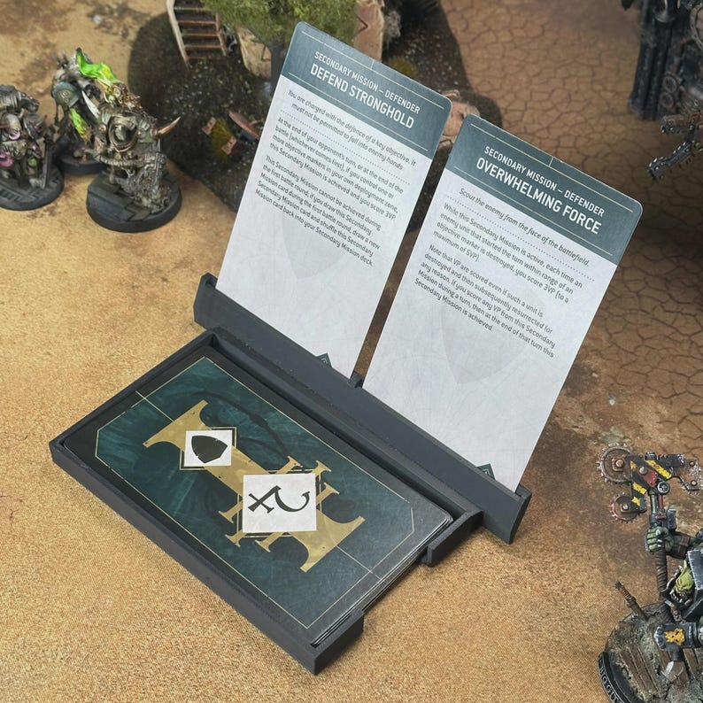 May include: A black plastic card holder displaying two vertical mission cards titled Defend Stronghold and Overwhelming Force, with a stack of dark green game cards featuring gold lettering and white symbols resting in the base tray.