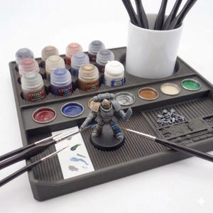 May include: A gray plastic painting station with various paint pots, brushes, and a miniature figurine. The station holds multiple paint colors, including red, blue, and gold. A white ceramic cup holds several paint brushes.