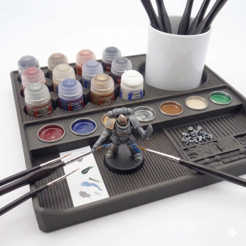 May include: A gray plastic painting station with various paint pots, brushes, and a miniature figurine. The station holds multiple paint colors, including red, blue, and gold. A white ceramic cup holds several paint brushes.