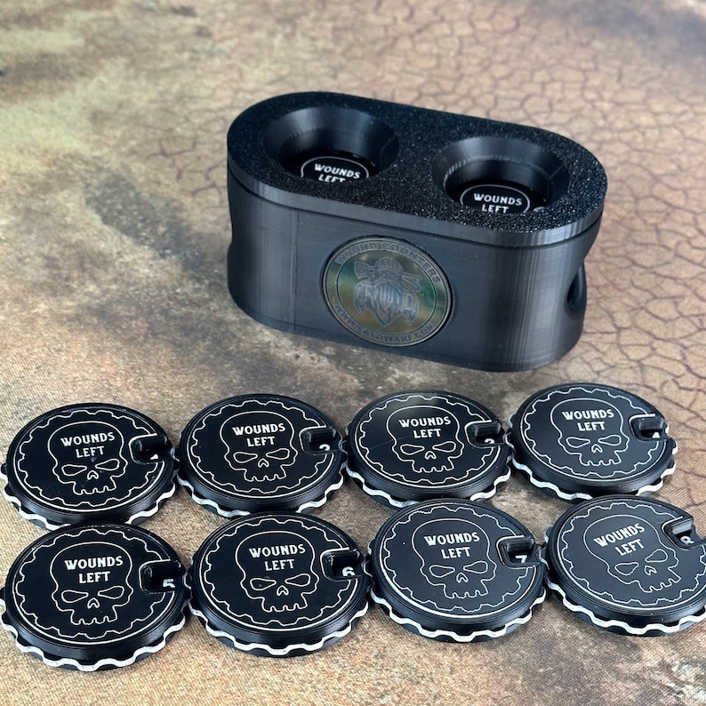 May include: Black plastic container with two compartments for holding small round tokens. Eight black tokens with white trim and a skull graphic are arranged around the container. Each token has the text "Wounds Left" printed on it.