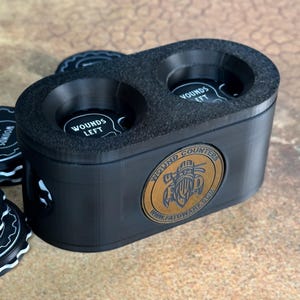 May include: Black wound counter with two circular compartments labeled "Wounds Left". The counter has a gold metal logo that reads "Wound Counters" and "www.fatdwarf.com".