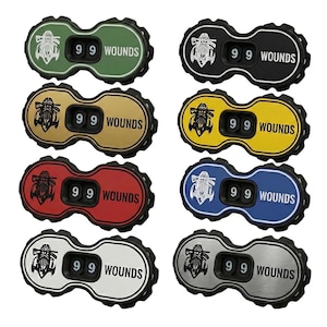 May include: A collection of eight bottle cap-shaped counters in various colors, each displaying the number "99" and the word "WOUNDS." The counters feature a stylized graphic and are in green, black, gold, yellow, red, blue, silver, and a combination of silver and black.