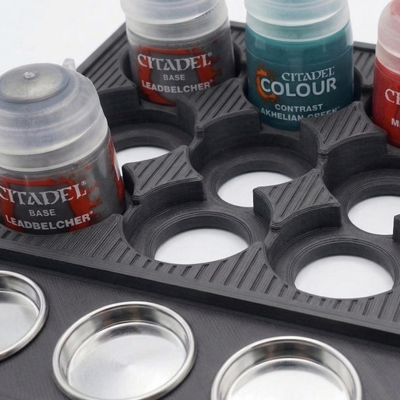 May include: A black paint bottle holder with multiple circular slots for paint bottles and small metal mixing pots. The holder contains Citadel brand paint bottles in various colors, including Leadbelcher and Akhelian Green.
