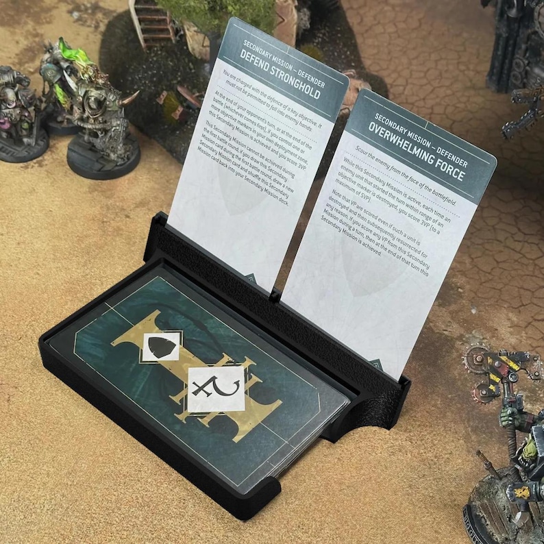 May include: A black plastic card holder displaying two upright mission cards titled Defend Stronghold and Overwhelming Force, with a stack of dark green game cards featuring gold lettering and white symbols resting in the base tray.