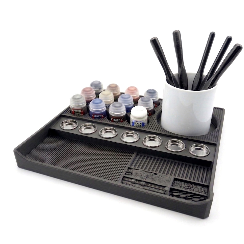 May include: A black paint station with a variety of Citadel paint pots, small metal paint wells, and a white ceramic cup holding black paint brushes. The paint station has a textured surface and a small compartment for tools.