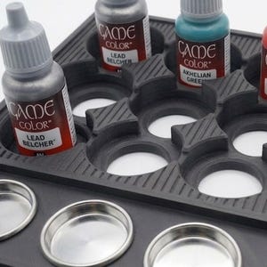 May include: A black paint bottle holder with multiple compartments, holding several bottles of Game Color acrylic paint. The bottles are labeled with paint names like "Lead Belcher" and "Akhelian Green". Several small, round metal containers are in the foreground.
